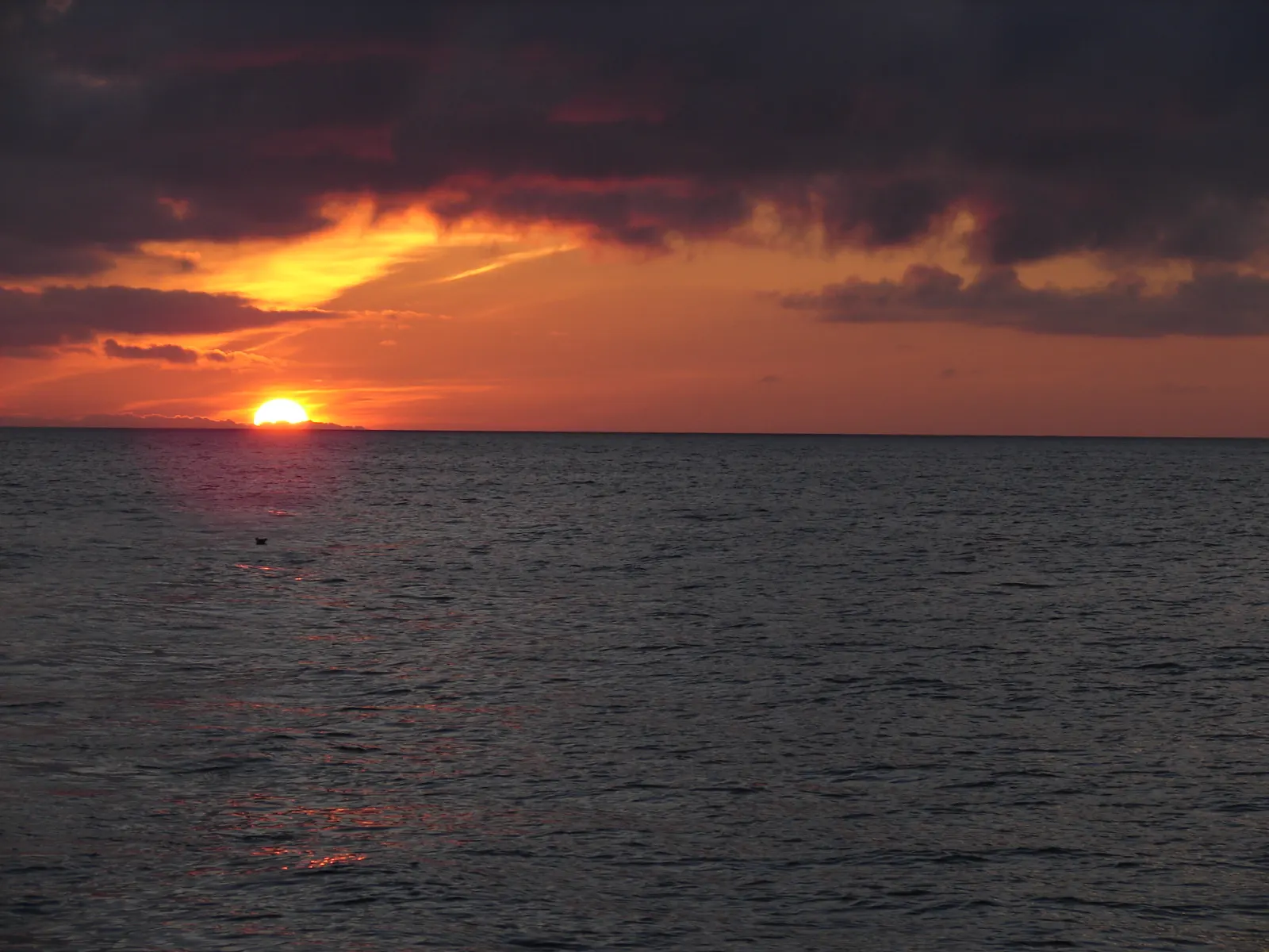 The late-day sun sinking to the ocean horizon beneath orange-lit clouds.