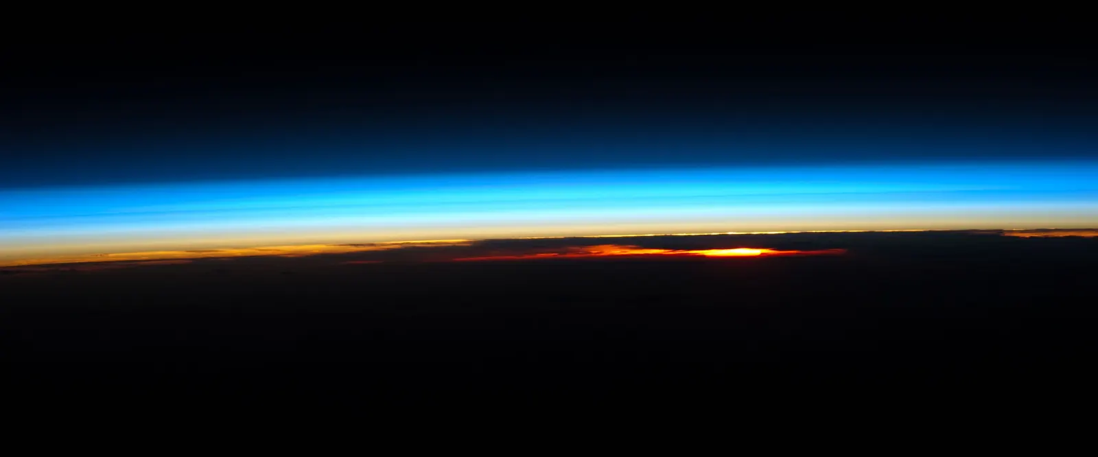 Earth's atmosphere seen edge-on from the International Space Station at sunset, with an orange glow at the bottom fading up through white into deep blue bands.